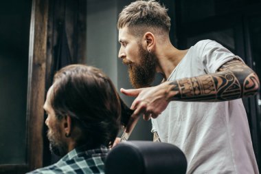 Calm attentive barber with long beard and tattoos on his arms touching long hair of his client and looking into the mirror
