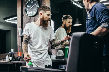 Uzun sakallı sakin profesyonel berber arkadaşının yanında duruyor ve bira içerken saç kesme bıçaklarını tartışıyor