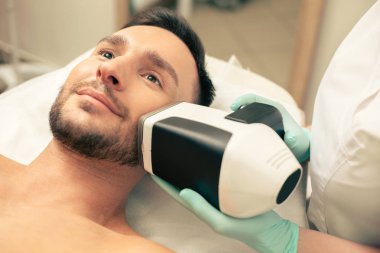 Genç sakallı adam yüzüne yakın modern bir cihaz yardımıyla cilt analizi prosedürü geçiyor