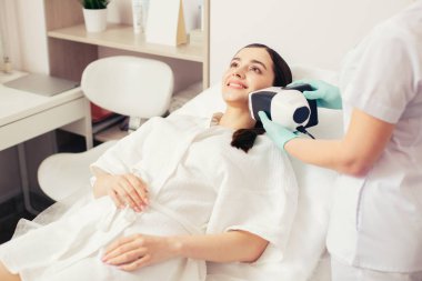 Uzun saçlı kadının bel kadar bir tıbbi kanepede yatan ve modern bir araç yardımıyla onun cilt durumu tam bir bakış sağlarken gülümseyen