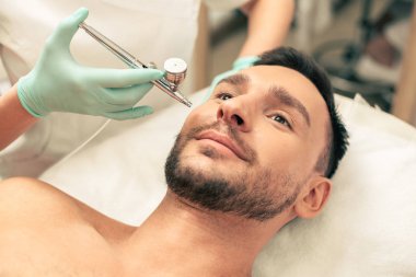 Genç sakallı adam yalan ve gülümseyen ise kauçuk eldiven oksijen spreyi tedavi prosedürü yürüten kozmetolojist