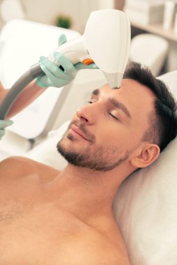 Sakallı genç adam kapalı gözlerle yastık üzerinde yatan ve prosedür sırasında alnında modern bir lazer cihazı tutan lastik eldiven el iken gülümseyerek