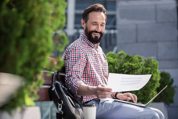 Work with pleasure. Cheerful handsome man sitting on the bench while using his laptop