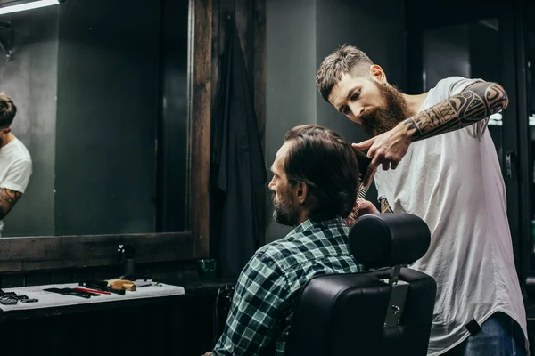 Calm attentive barber standing with comp in his hand and cutting long hair of young man