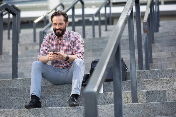 Urban style of life. Cheerful pleasant man using his phone while sitting on the stairs outdoors