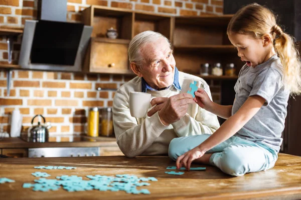 Tjis peace. Nice little girl sitting on the table while assembling jigsaw puzzle with her grandfather