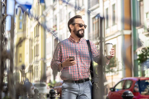 Urban dweller. Cheerful bearded man drinking coffee while walking in the city