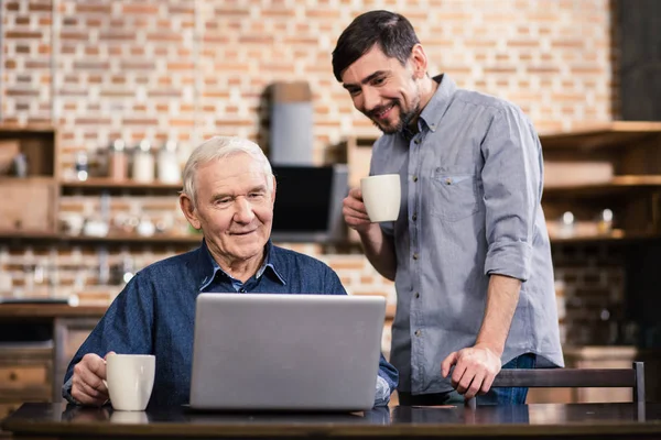 Modern user. Positive elderly man using his laptop while his son standing nearby
