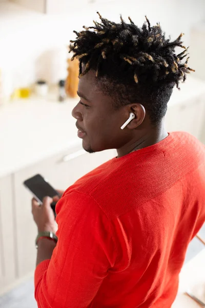 Young man standing alone and turning his head while using modern smartphone with wireless earphones and looking away