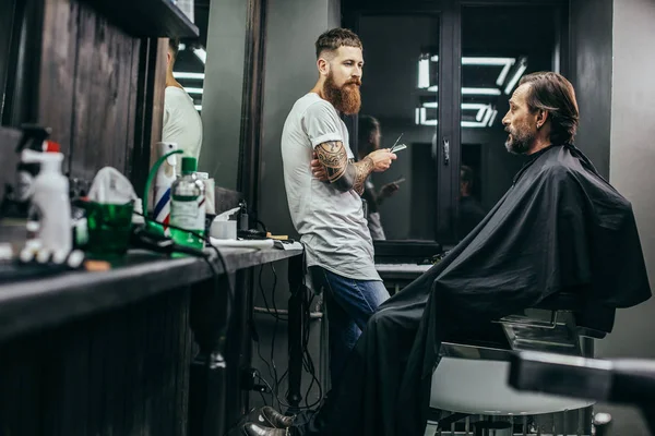Calm bearded man covered in hairdressing gown and talking to his professional barber