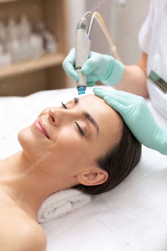 Close up of calm relaxed lady lying with her eyes closed and professional cosmetologist cleaning her skin with dermabrasion tool