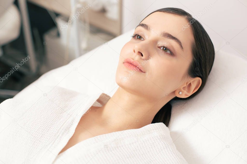 Face of a young dark haired woman visiting a modern clinic and staying calm while lying on a medical couch in white bathrobe