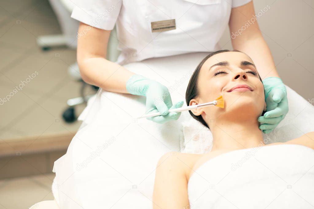 Peaceful young lady lying with her eyes closed and smiling while a beautician cleaning her face with a brush