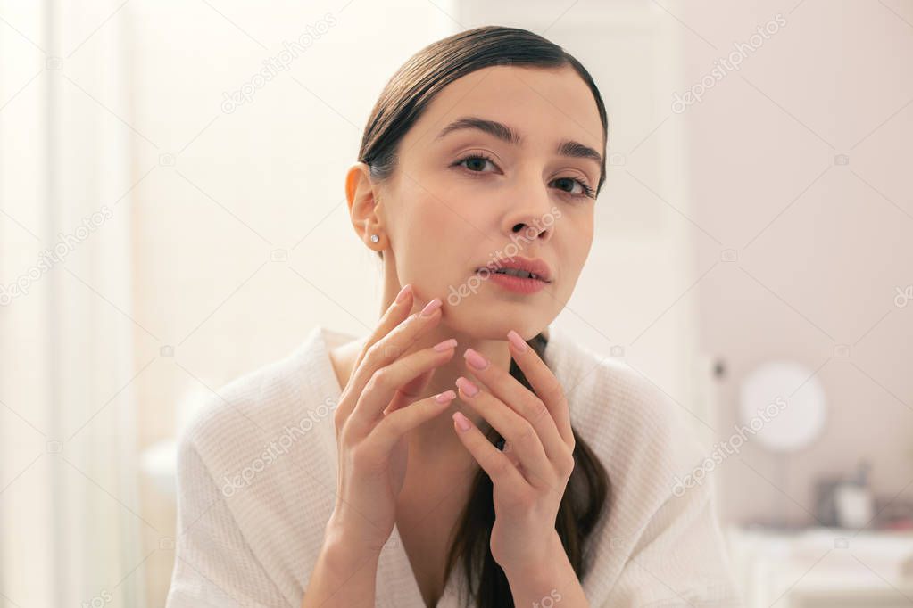 Calm young woman in a white towel looking concentrated while touching her chin and looking at the skin condition