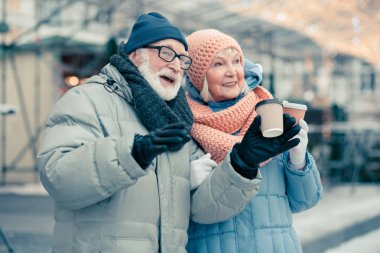 Kış giysileri rahat yaşlı çift ellerinde kahve karton bardak ile açık havada vakit geçirmek ve bir gülümseme ile uzağa bakıyor