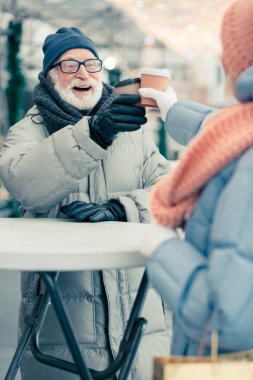 Güzel kış lık güzel elbiseli mutlu yaşlı adam, yuvarlak sokak masasının yanında açık havada durup eşiyle kağıt kahve fincanı yaparken gülümsüyor.