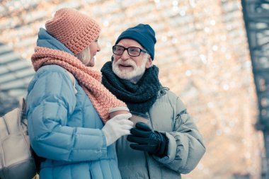 Rahat yaşlı çift kış lık kıyafetlerle birlikte ayakta durup konuşuyorlar. Arka planda Noel aydınlatma