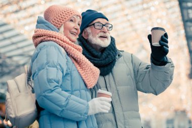 Küçük sırt çantası ile yaşlı bayan onun yaşlı kocasına yakın duran ve onunla gitmek için kahve keyfi yaparken gülümseyerek. Arka planda Noel aydınlatma