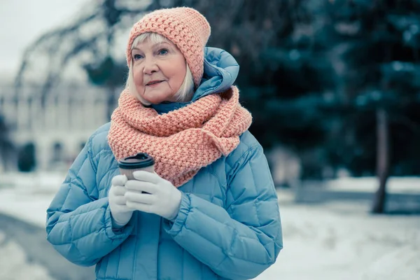 Waist up of a peaceful old lady in warm clothes standing outdoors in a winter day and looking away while holding a carton cup of coffee
