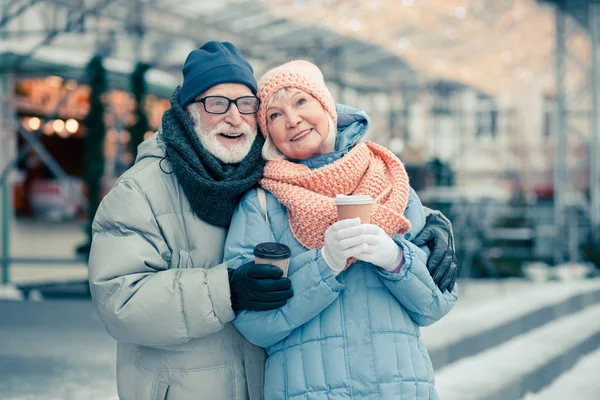 Street with Christmas illumination on the background. Elderly couple smiling while hugging and holding carton cups of coffee