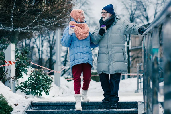 Elderly people in winter clothes holding cups of hot chocolate and looking at each other with a smile. Man touching handrail while walking downstairs