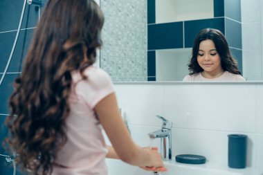 Sakin, güzel bir kızın aynadaki yansıması. Gülümseyen ve ellerini su ve sabunla yıkarken gözlerine bakan.