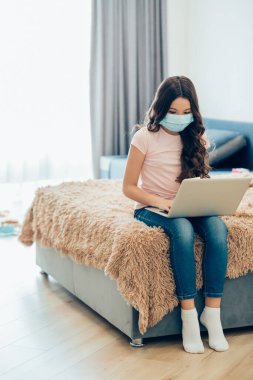 Kucağında modern bir dizüstü bilgisayarla yatakta yatıp ekranına bakan uzun saçlı sakin bir kız. Yüzünde tıbbi maske var.