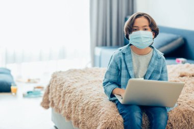 Yatağının kenarında otururken kucağında dizüstü bilgisayar tutan ve yüzünde tıbbi maske olan sıradan giysili bir çocuk.