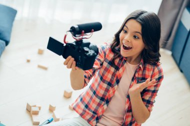Cute happy girl gesturing and smiling while leading a video blog and recording herself on a camera clipart
