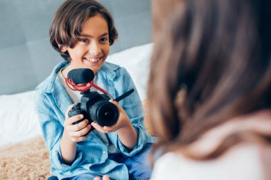 Neşeli hevesli çocuk elinde mikrofonla bir kamerayla önünde oturan arkadaşına gülümsüyor.