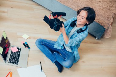 Gladsome çocuğu elinde kamera ve mikrofonla yerde oturuyor ve video çekiyor. Modern dizüstü bilgisayar ve önünde notlar.