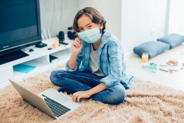 Laptop ekranının önünde yatağında otururken tıbbi maske takan rahat bir çocuk.