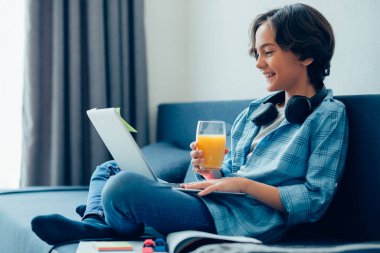 Kanepede oturan pozitif çocuk kucağında modern bir dizüstü bilgisayarla ekrana bakarken elinde bir bardak portakal suyu tutuyor.