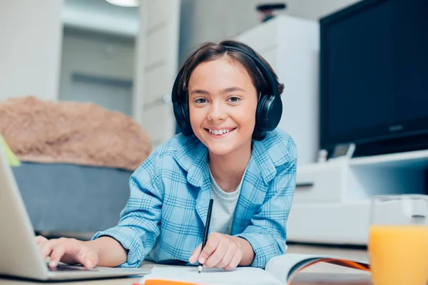 积极的男孩 头戴耳机 手握铅笔 躺在笔记本电脑屏幕前的地板上 — 图库照片
