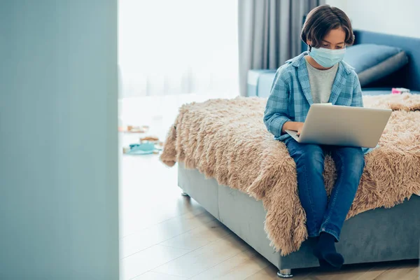 Boy in medical mask sitting on the bed with a laptop on his laps. Template banner