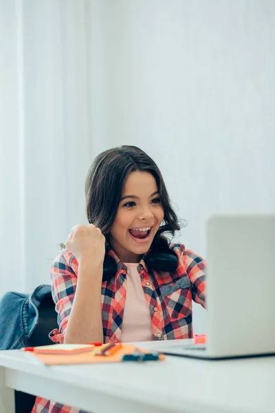 可爱的姑娘坐在桌旁 面前拿着笔记本电脑 微笑着看着屏幕 庆祝胜利 — 图库照片