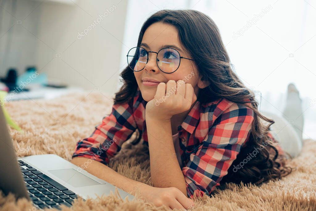 Mirthful pretty girl wearing glasses putting her chin on the hand and smiling while lying in front of a laptop