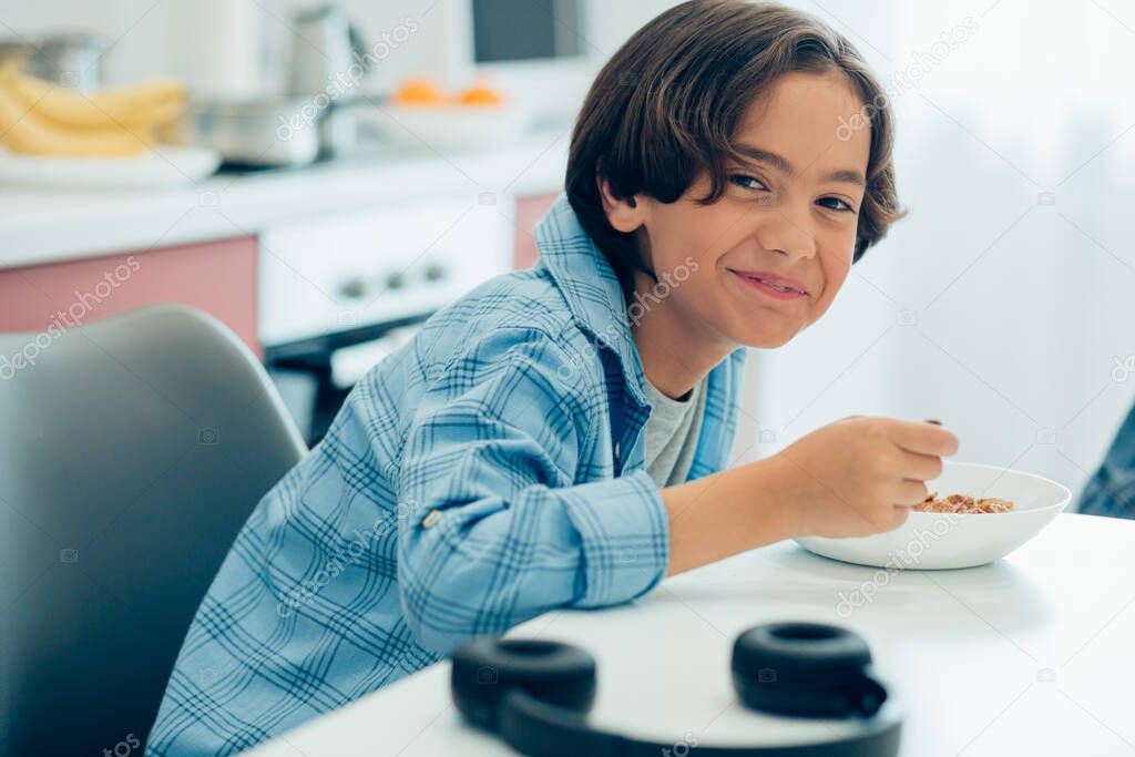 Positive schoolboy looking happy while sitting at the table at home with a bowl of cornflakes and a spoon in his hand