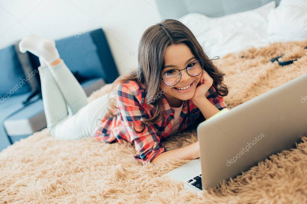 Joyful girl wearing glasses and looking at the laptop screen while lying on the bed with her feet up