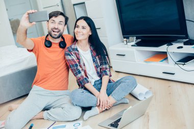 Neşeli genç bir adam güzel bir bayanla oturuyor ve onunla fotoğraf çekerken elinde bir akıllı telefon tutuyor. Dizüstü bilgisayar ve çizelgeler yere.