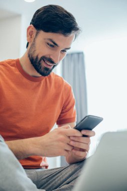 Elinde modern bir akıllı telefonla evde oturan neşeli, genç bir yetişkin ve ekranına bakıyor.