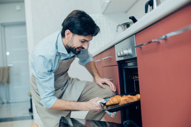 Mutfakta tek dizinin üstünde duran ve kruvasanlara bakıp gülümseyen mutlu bir adam.