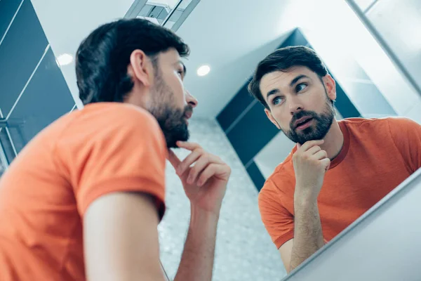 鏡の前に立って 反射を見ながら顎に触れる好奇心旺盛な若い男 — ストック写真