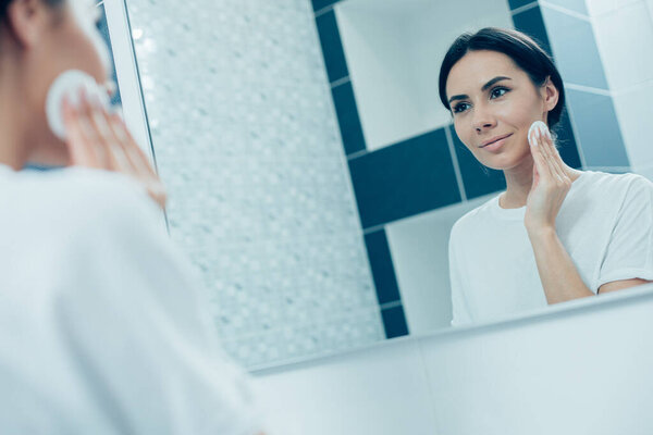 Contented young woman looking at her reflection in the mirror and smiling while touching her cheek with a cotton pad