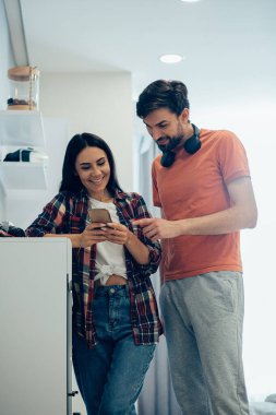 Gülümseyen bir kadının yanında duran ve elindeki akıllı telefonun ekranına bakan olumlu genç adam.