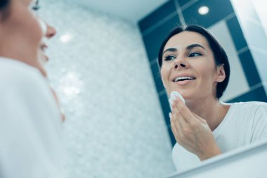 Pamuk yastıkla çenesine dokunan ve banyo aynasında yansımasına gülümseyen güzel bir kadın.
