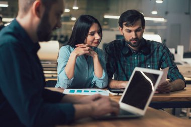 Ön planda iki kişi grafik çizelgeleriyle oturup onlara bakarken dizüstü bilgisayarda çalışan adam.