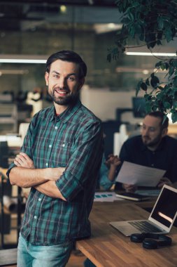 Pozitif çekici genç adam sırtı iş arkadaşlarına dönük duruyor ve gülümserken kollarını kavuşturuyor.