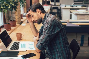 Genç adam masanın üzerinde grafik çizelgeleri ile otururken alnına dokunuyor ve modern bir dizüstü bilgisayarın ekranına bakıyor.