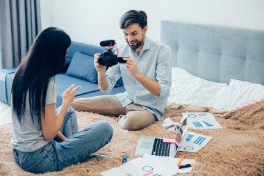 Yatakta oturan heyecanlı genç adam kameraya bayan meslektaşını kaydediyor. Modern dizüstü bilgisayar ve grafik çizelgeleri.